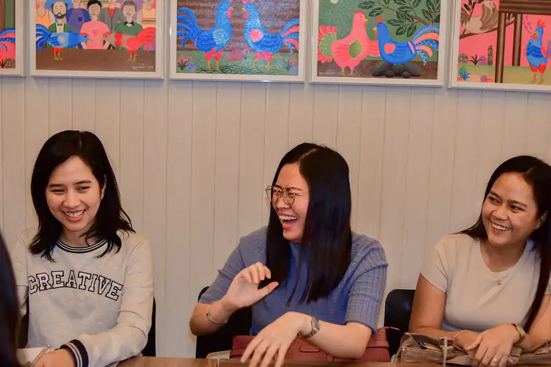 Three young Filipino women laughing together at a table in a colourfully decorated room