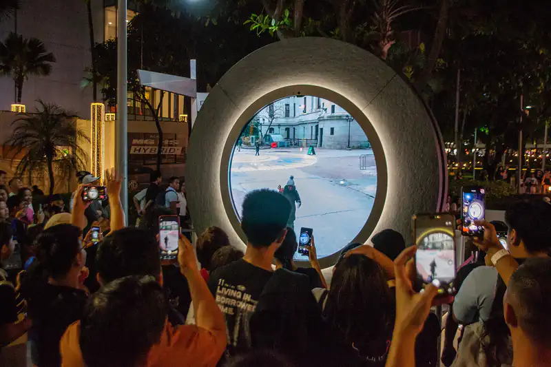 A night crowd in Metro Manila raising smartphones to photograph a lit-up circular portal sculpture
