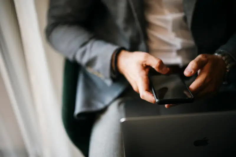 Close-up of a person’s hands holding a smartphone while sitting with a laptop on their lap