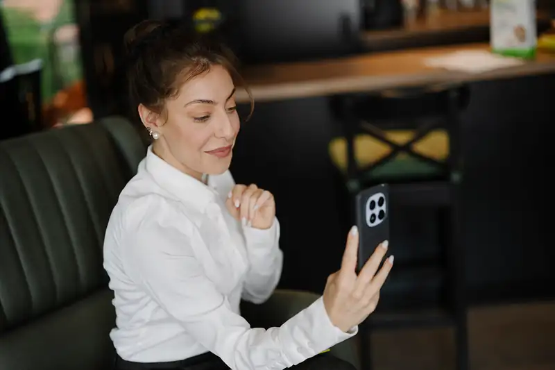A woman in a white blouse sitting in a café chair holding up her smartphone for a video call